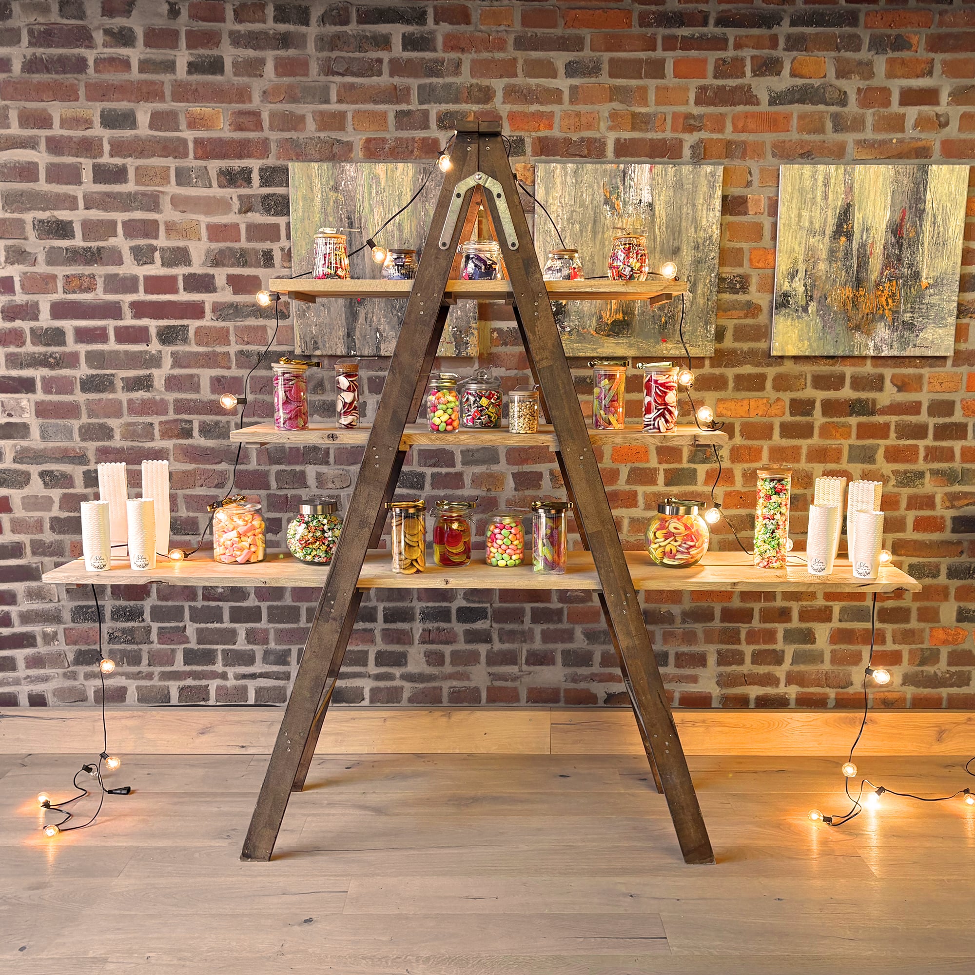 Dreistufige Candybar mit verschiedenen Bonbongläsern auf Holzregalen, die an einer Holzleiter vor einer Backsteinwand hängen, umrahmt von Lichterketten