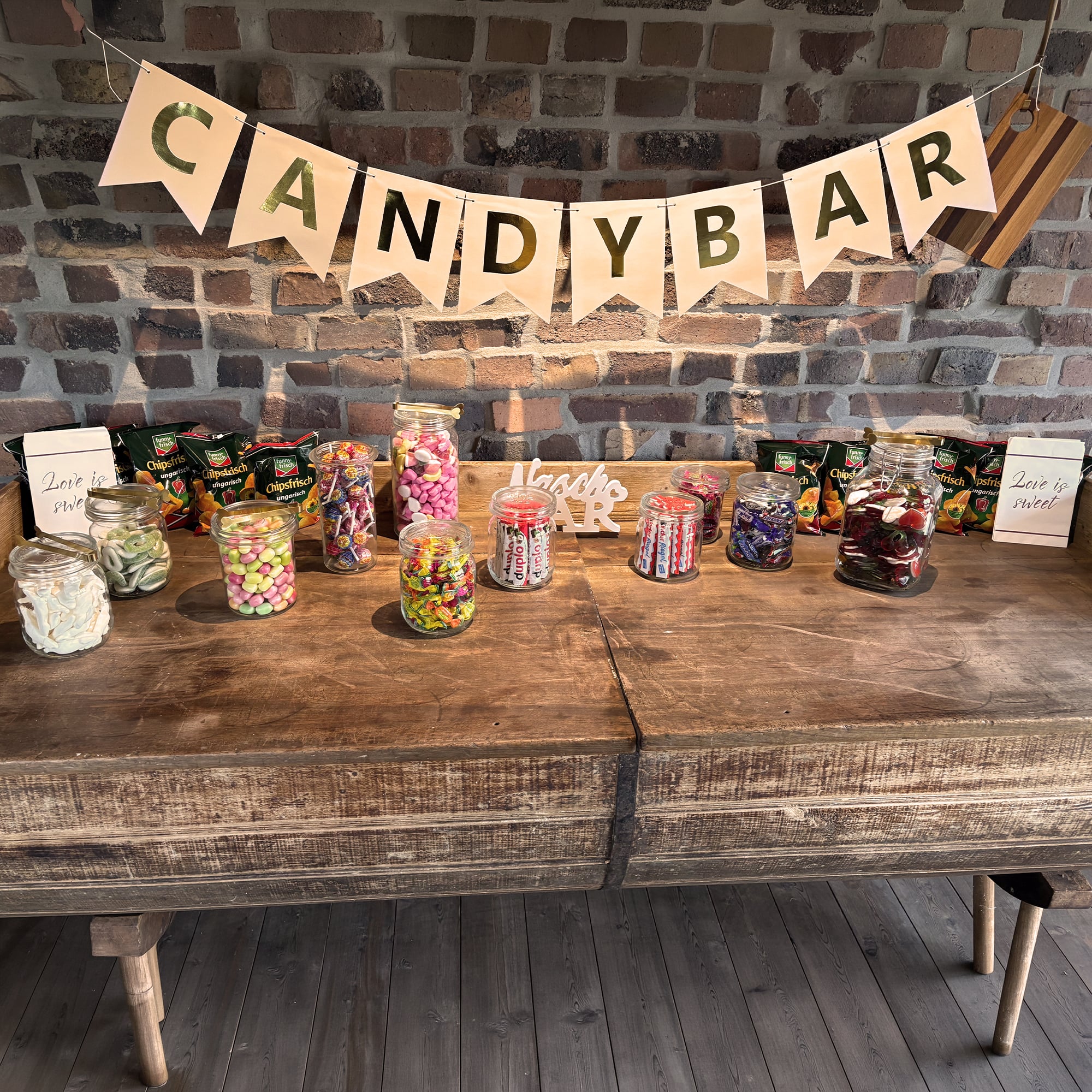 Mehrere Gläser voller Süßigkeiten als Candybar für Hochzeit vor Backsteinwand mit Wimpeln auf denen das Wort "Candybar" steht