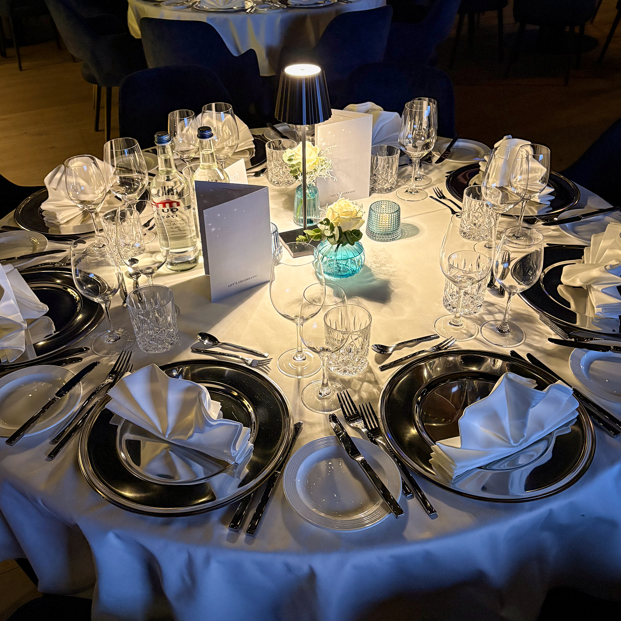 Runder festlich gedeckter Tisch mit weißen Servietten, Gläsern, Silberbesteck, kleinen Blumenarrangements und einer Tischlampe in einem dunklen Raum