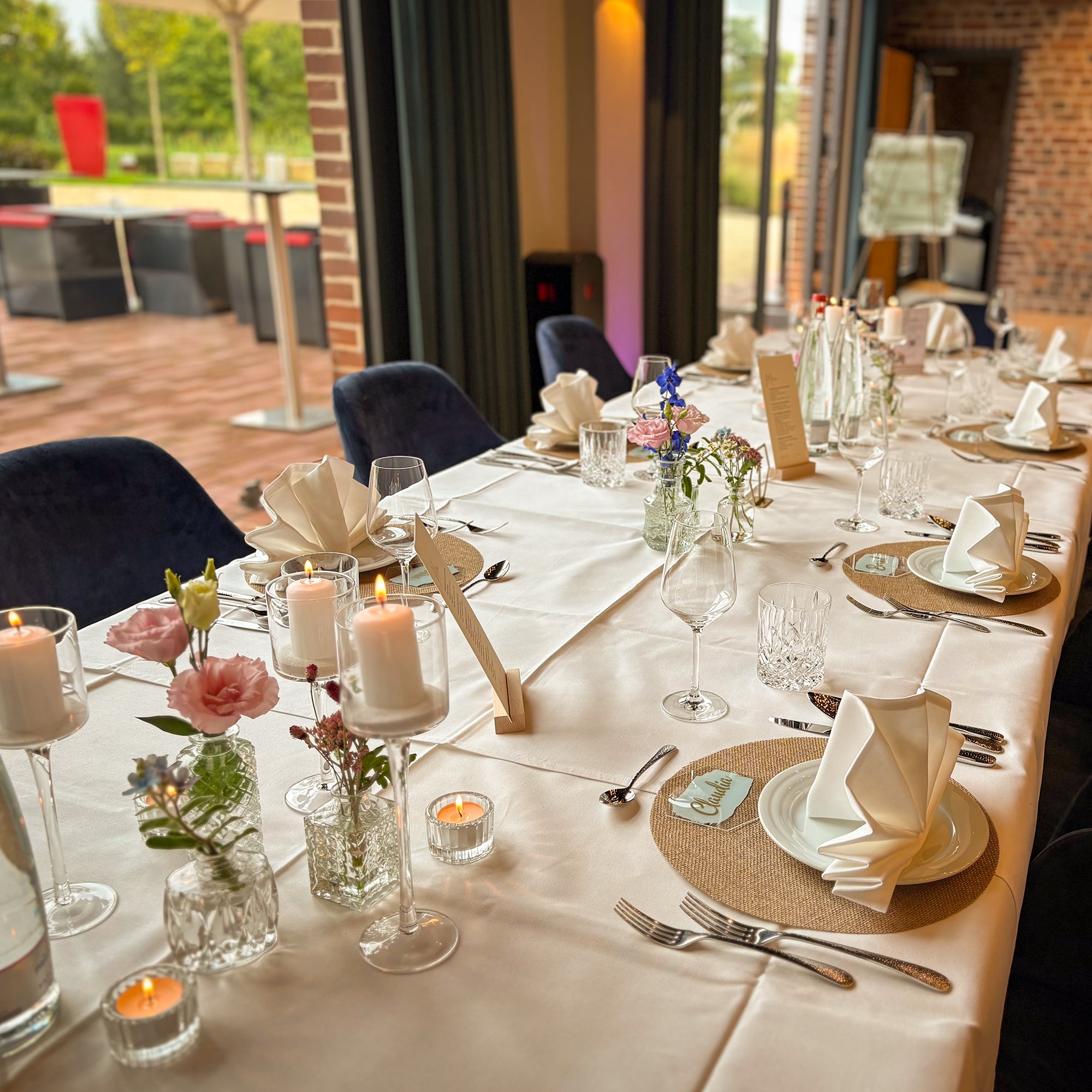 Festlich gedeckter Tisch mit weißen Servietten, Kerzen in Gläsern und kleinen Blumenarrangements in verschiedenen Vasen in einem hellen Raum mit Blick auf eine Terrasse.
