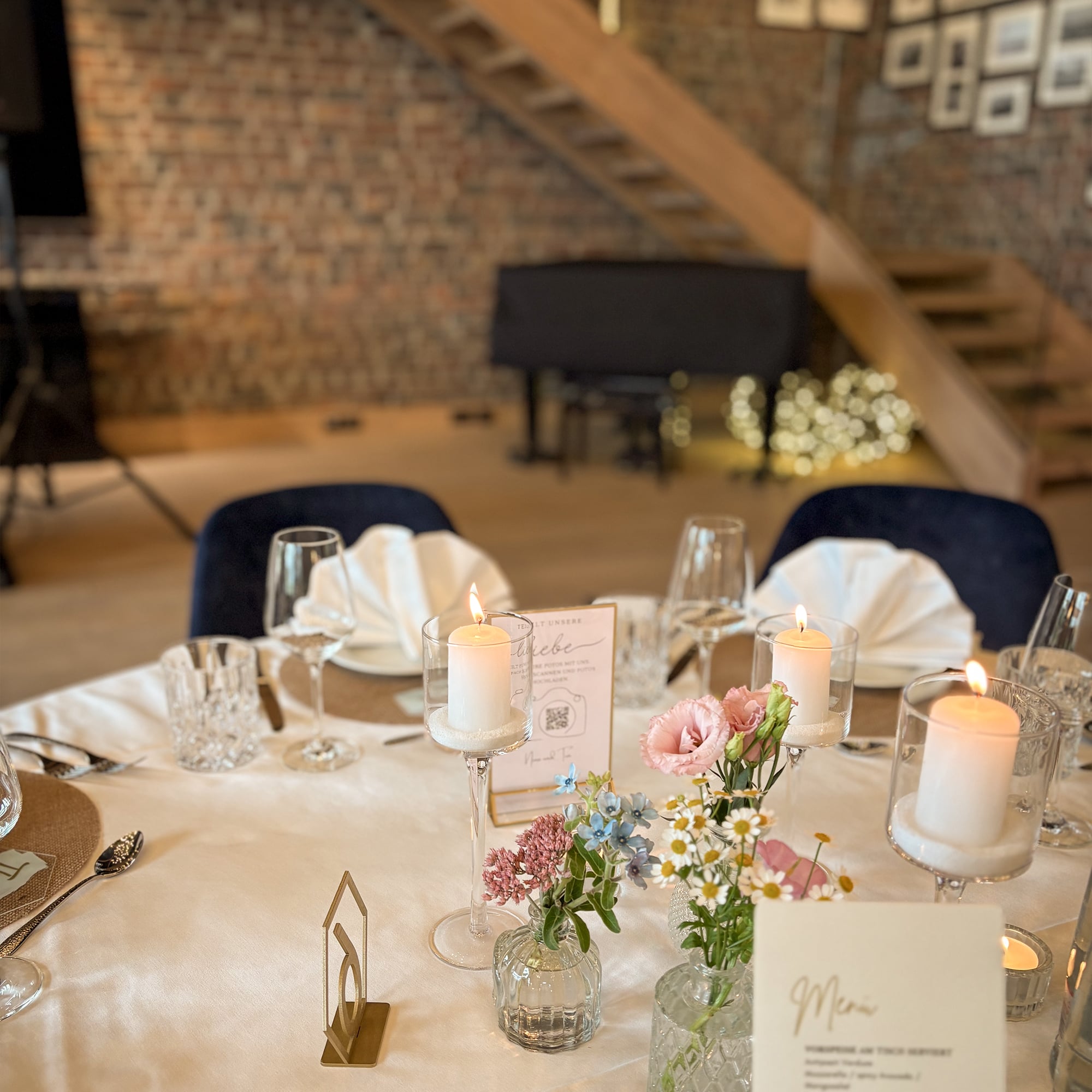 Festlich gedeckter Hochzeitstisch mit weißen Kerzen, kleinen Blumenvasen, gefalteten Servietten und Menükarte vor einer Backsteinwand mit Treppe und Klavier im Hintergrund