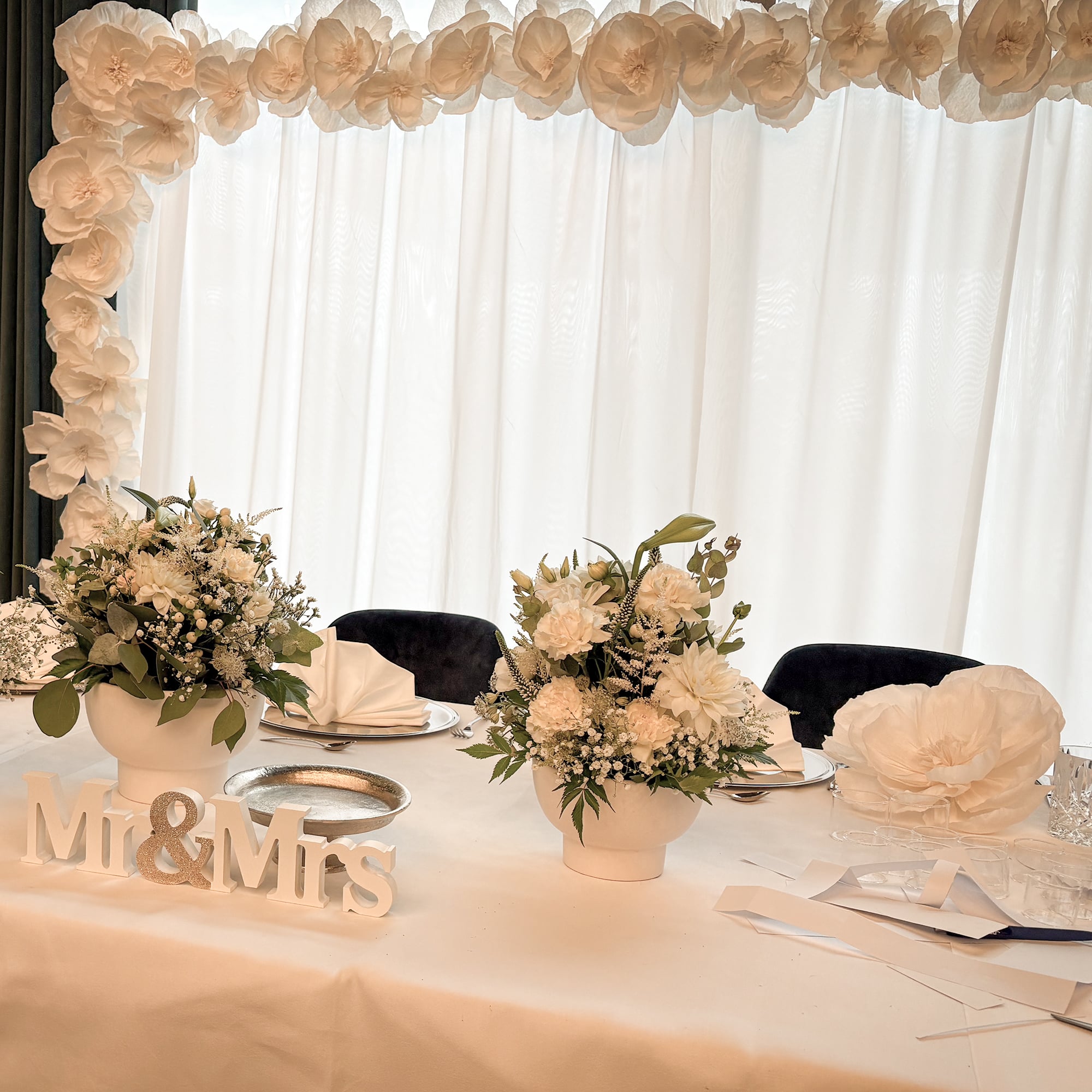 Hochzeitstisch mit weißen Blumenarrangements, Servietten, silbernem Teller und Schriftzug 'Mr & Mrs'